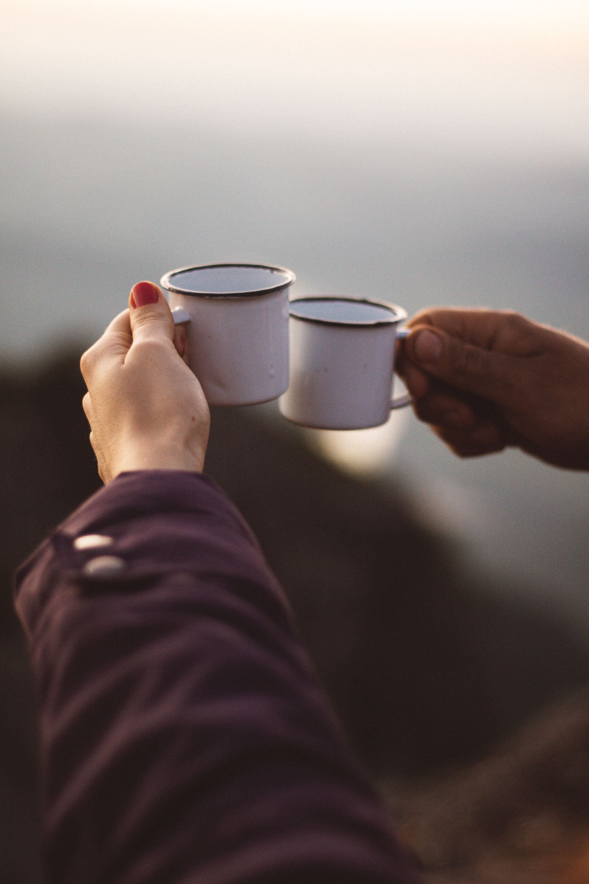 Two people toasting with white metal cups outdoors during sunset.