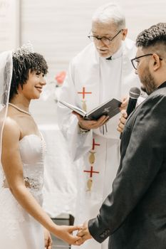 Bride and groom holding hands during a wedding ceremony with a priest officiating.