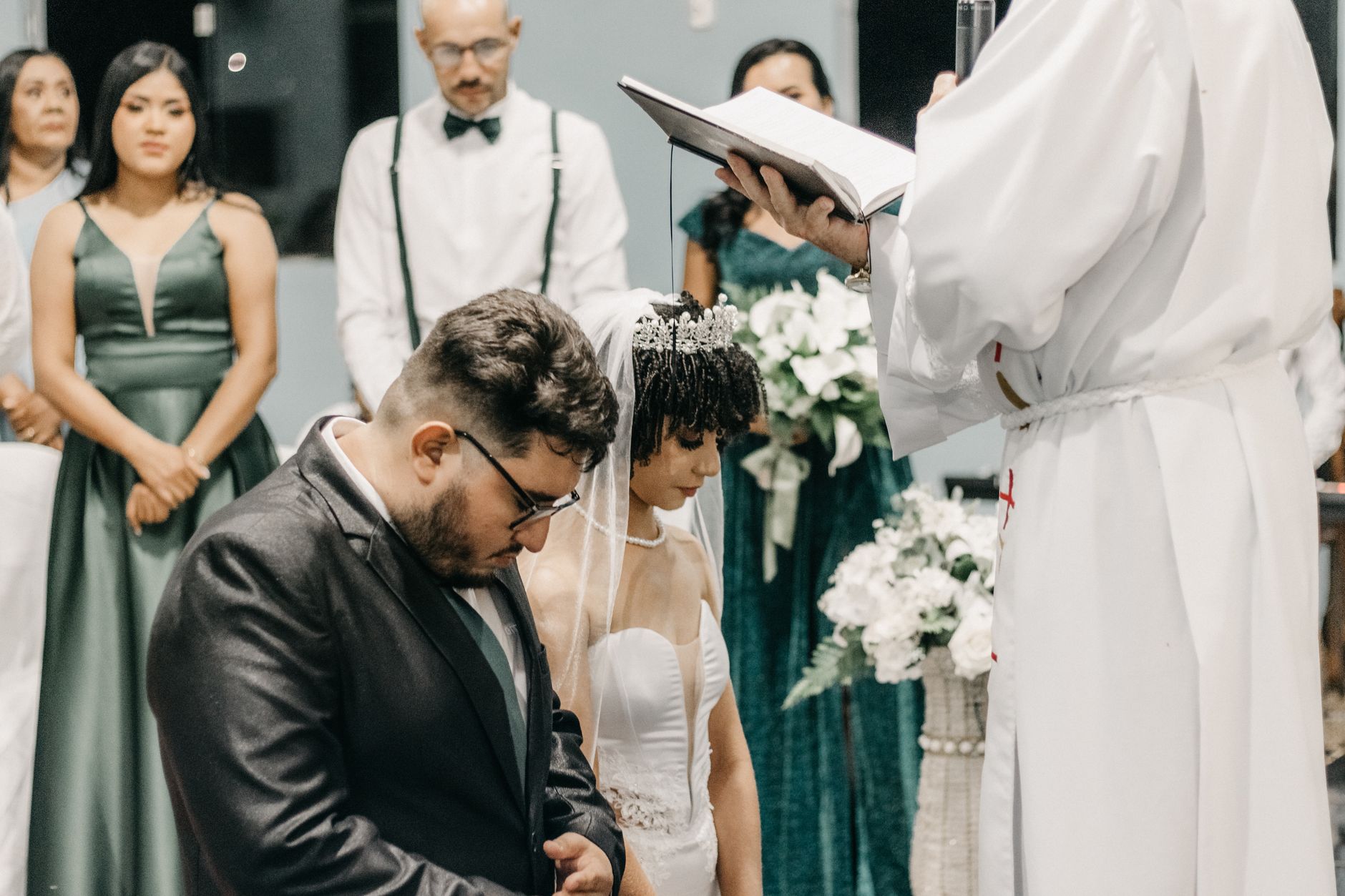 Komitmen Standar Pelayanan dan Dokumentasi Acara Pernikahan Bersama Jagarasa 1 An elegant indoor wedding ceremony with a bride and groom kneeling before a priest, surrounded by bridesmaids.