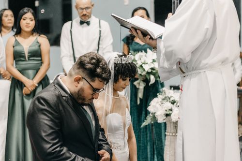 Free An elegant indoor wedding ceremony with a bride and groom kneeling before a priest, surrounded by bridesmaids. Stock Photo