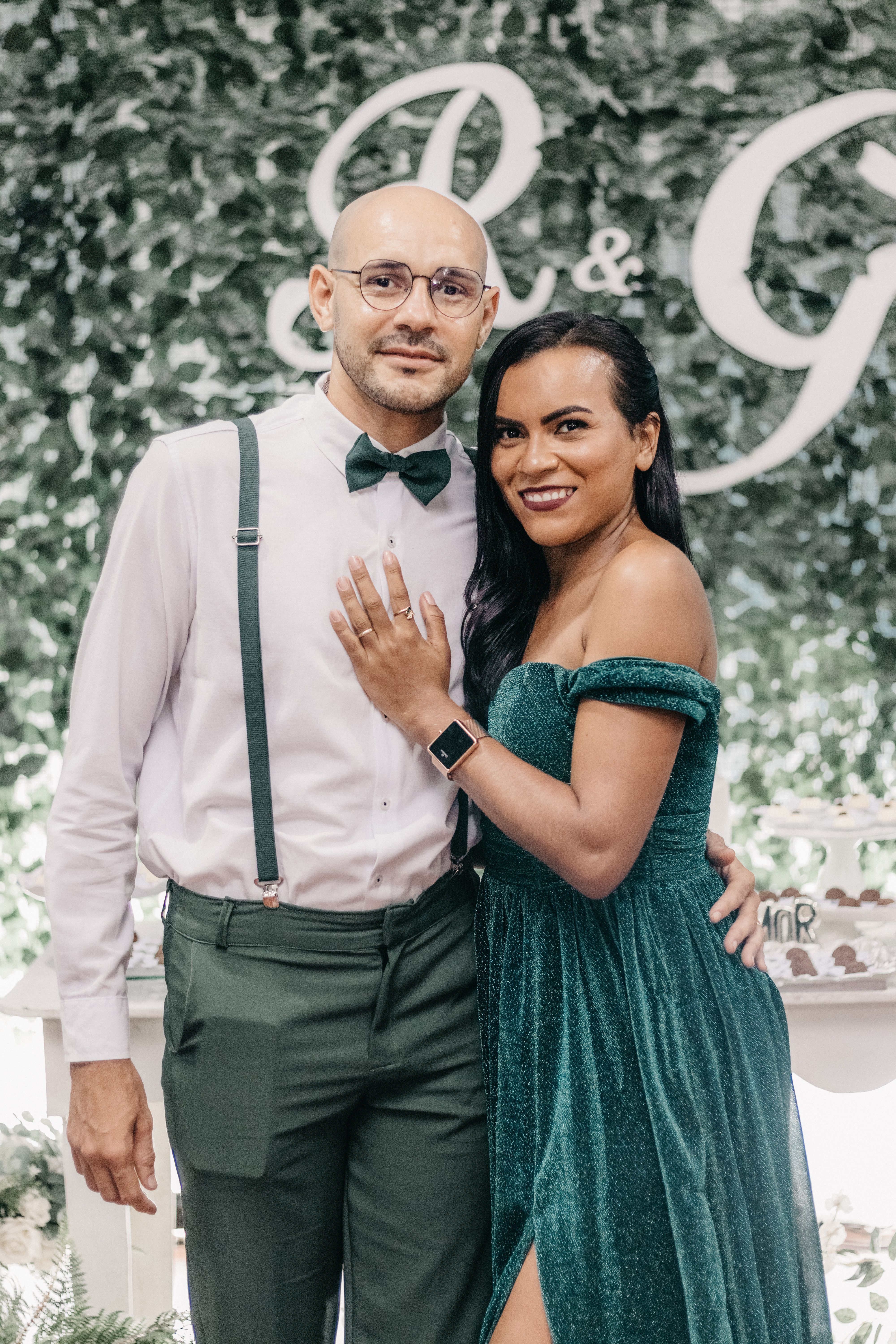 Happy couple elegantly posing indoors in formal attire, surrounded by lush greenery and initials backdrop.