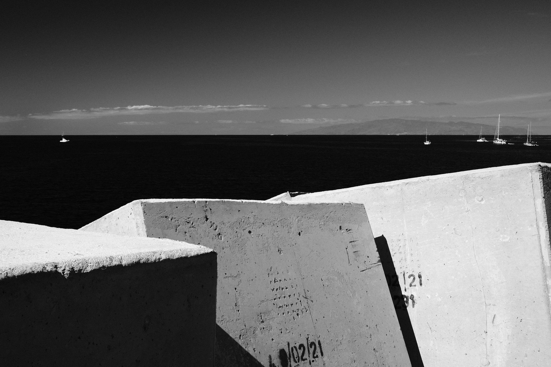 Monochrome view of concrete blocks by the ocean in Spain with boats in the distance.