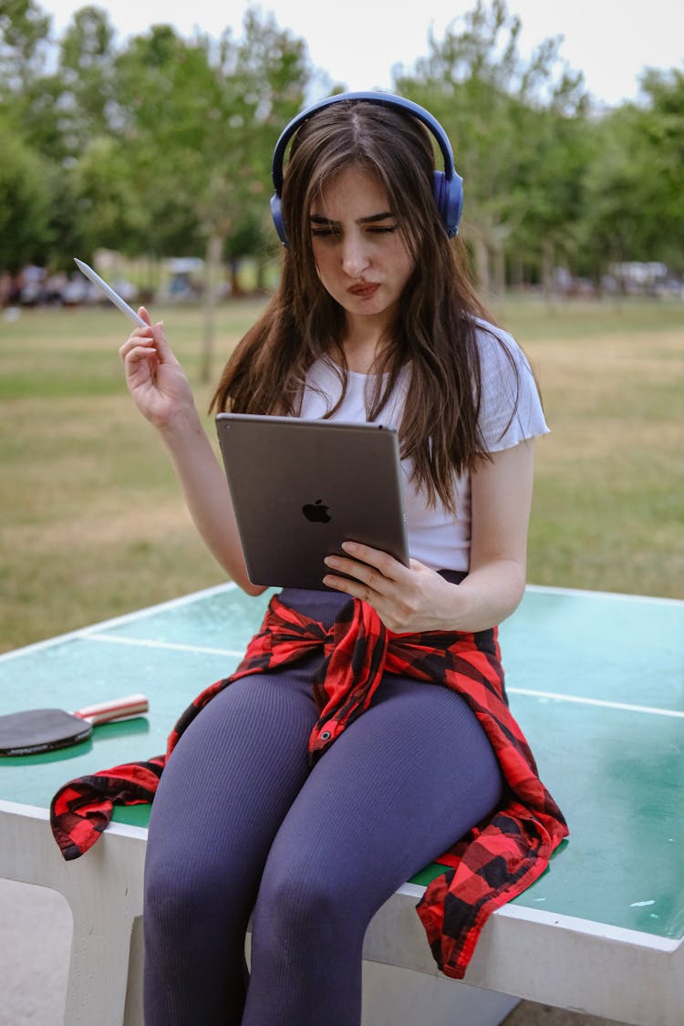 Young Woman Sitting On Table In Park With Ipad In Hand