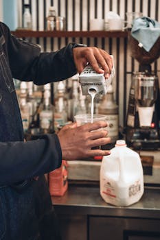 A barista skillfully pours milk from a carton into a coffee cup in a bustling cafe setting.