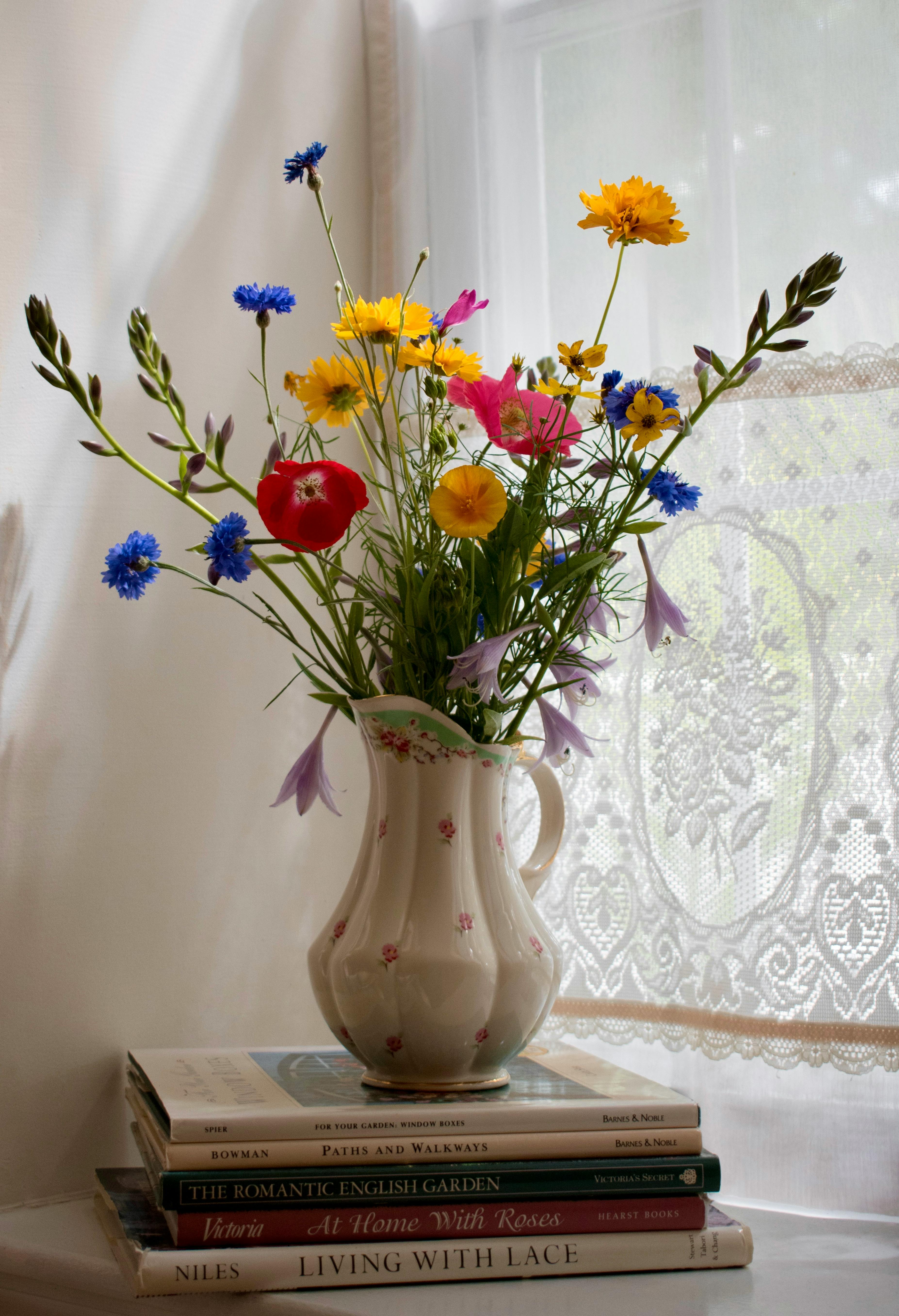 Vibrant wildflowers arranged in a vintage floral vase atop books by a lace curtain.