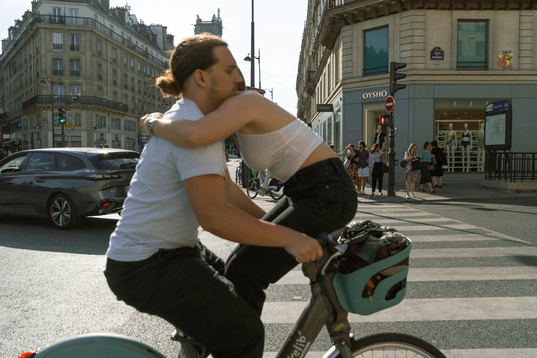 A couple hugging while cycling on a bicycle across a city crosswalk on a sunny day.