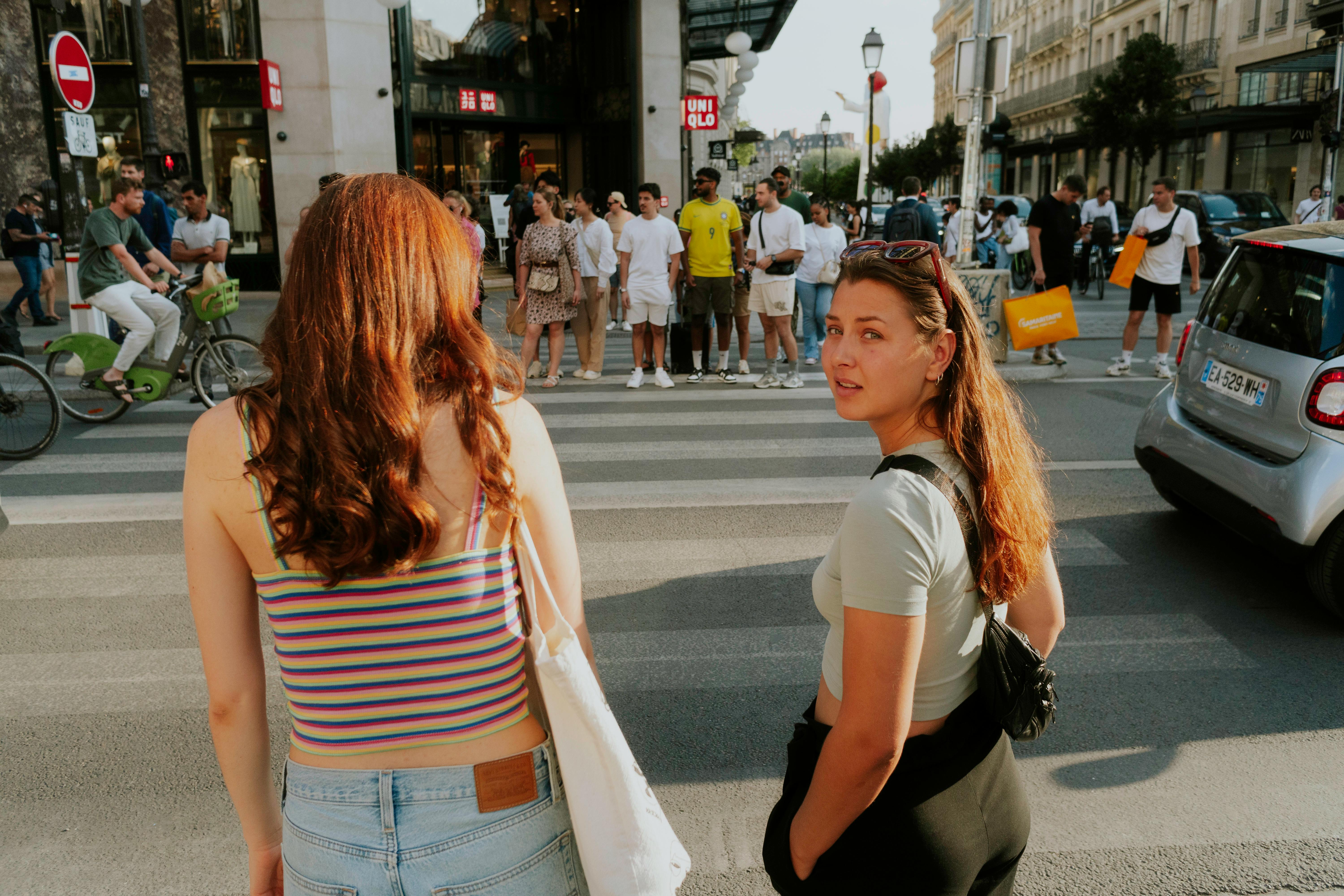 Two young women navigate a busy urban crosswalk, surrounded by city life and pedestrians.