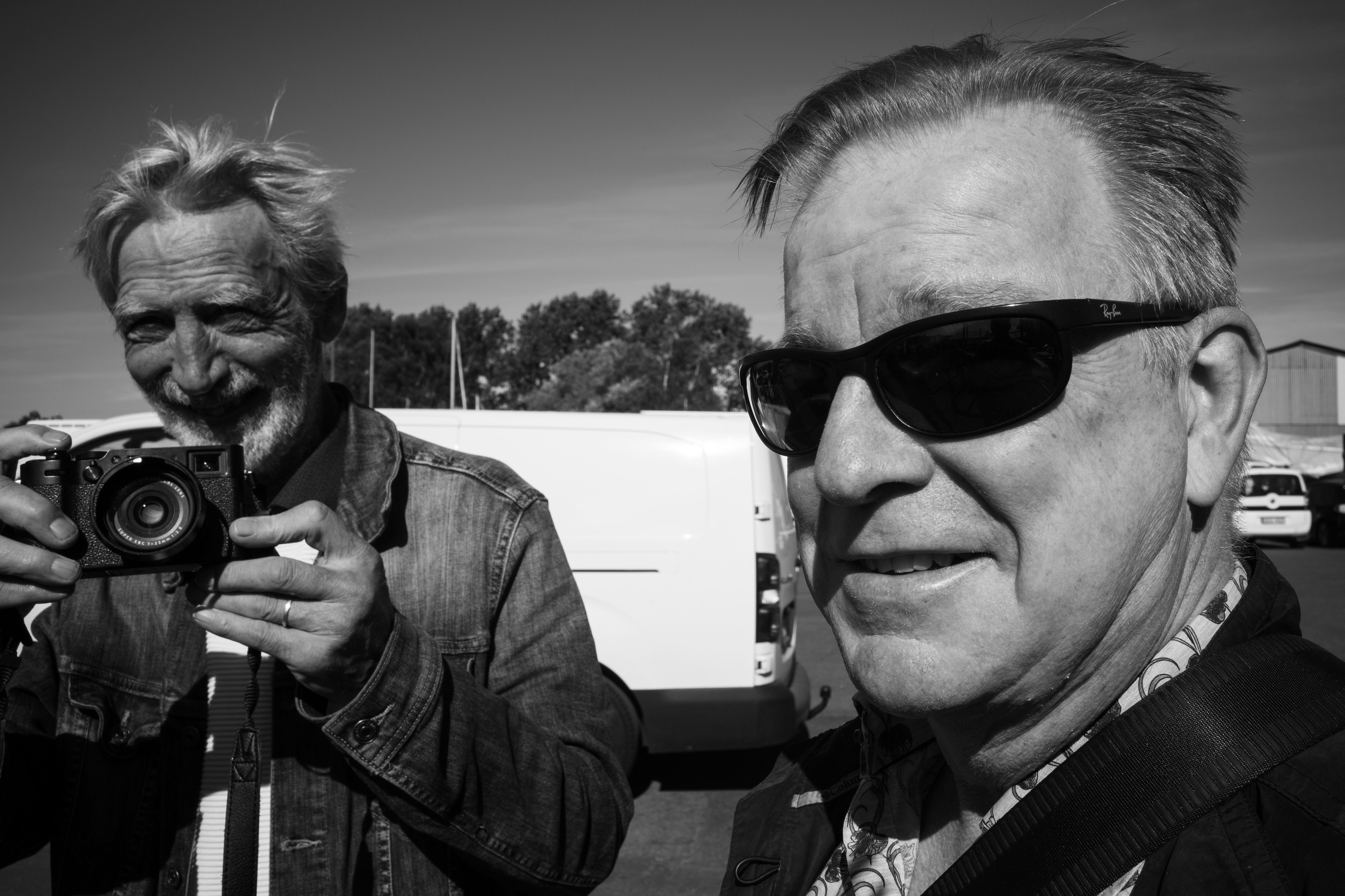 Elderly men outdoors, one holding a camera, both smiling in sunlight.