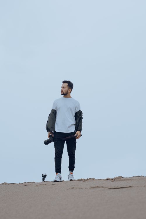 Free A man stands on a sandy hill holding a camera, dressed casually in a white t-shirt with clear skies overhead. Stock Photo
