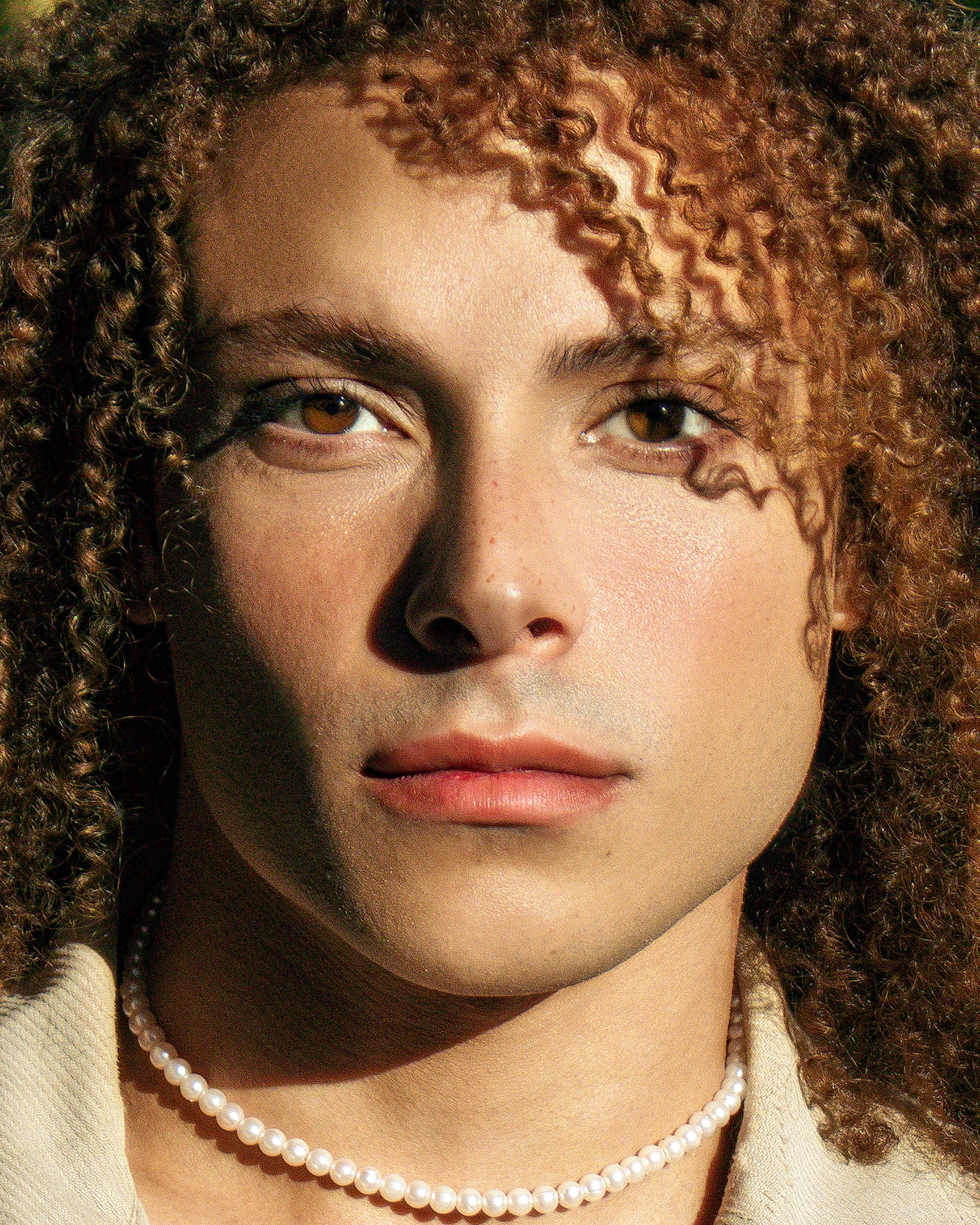 Close-up portrait of person with curly hair and pearl necklace in sunlight. Captivating expression.