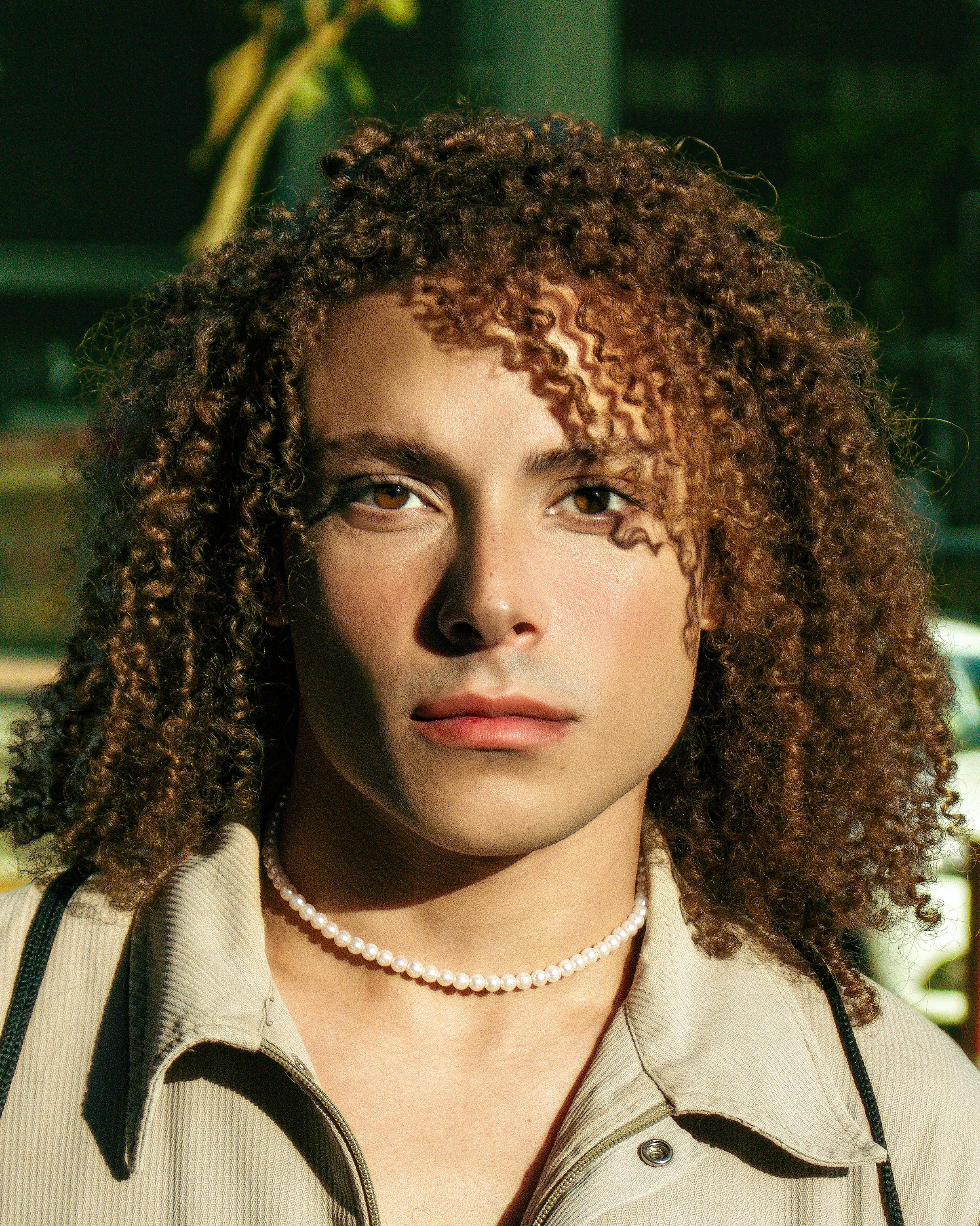 Close-up portrait of a young man with curly hair against a sunlit urban background.