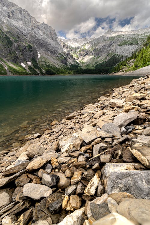 View of a Rocky Beach by a Lake in a Valley with Snowcapped Mountains in the Background 