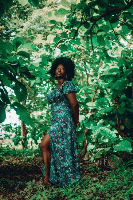 A woman gracefully poses in a floral dress surrounded by vibrant foliage in an outdoor setting.