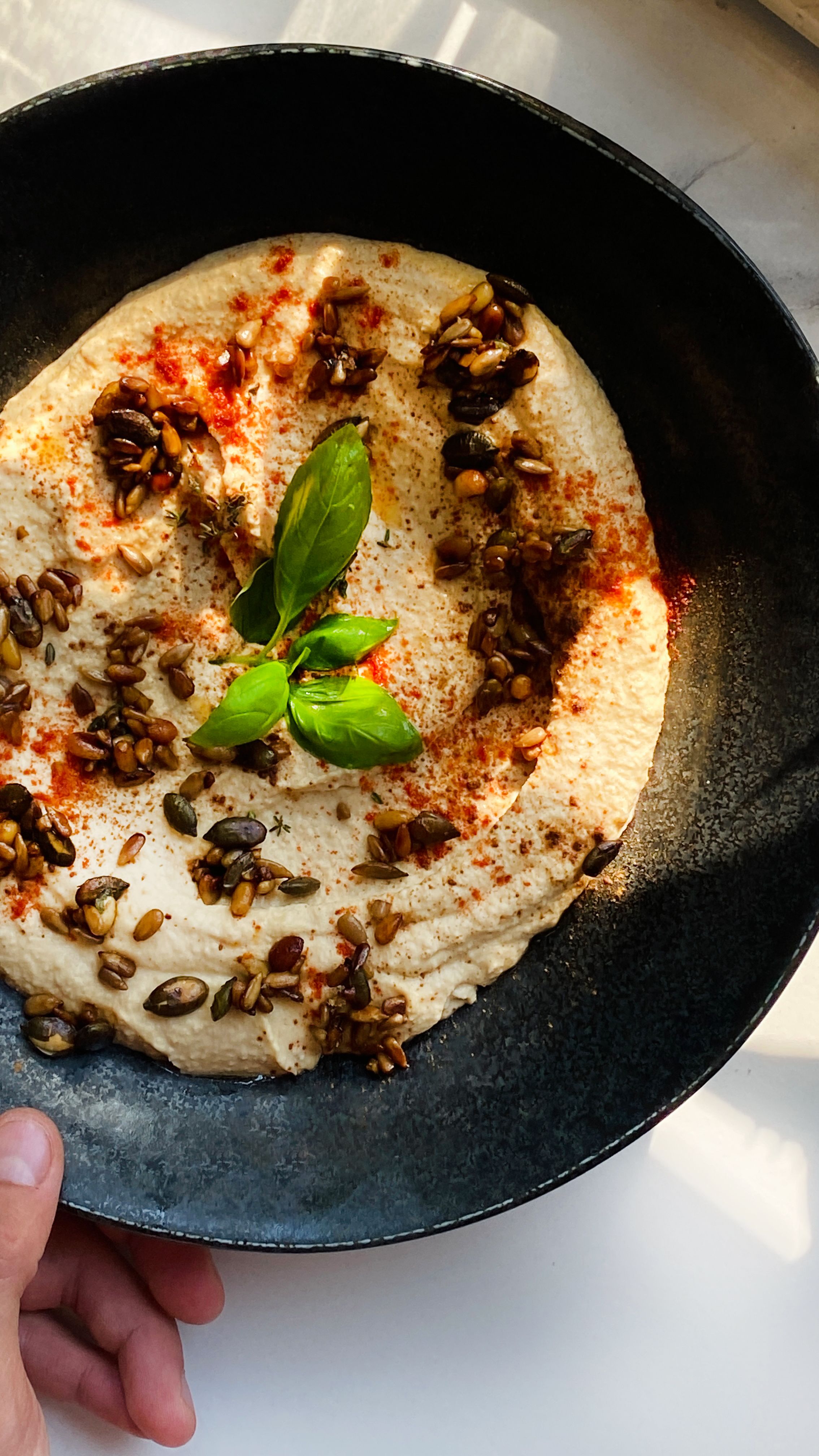 Delicious hummus garnished with seeds, spices, and fresh basil leaves in sunlight.
