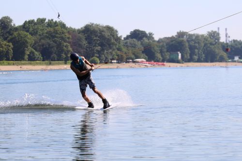 Free Man kitesurfing energetically on a calm lake during a sunny day, surrounded by lush greenery. Stock Photo