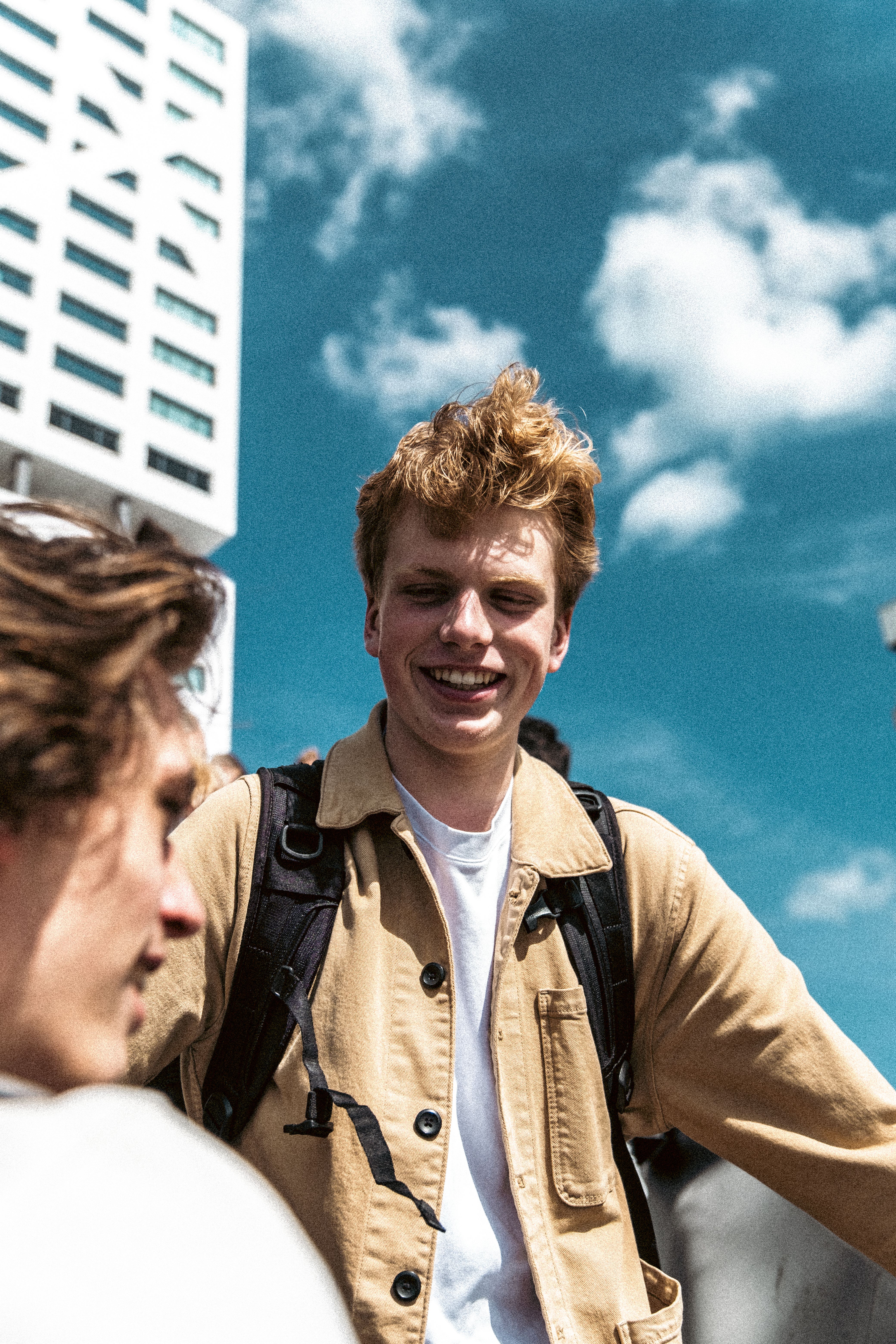 A cheerful young man wearing a beige jacket enjoys a sunny day in a city environment.