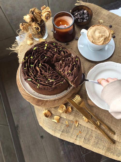 Woman Taking a Slice of a Chocolate Cake Standing on a Table next to a Coffee and a Candle