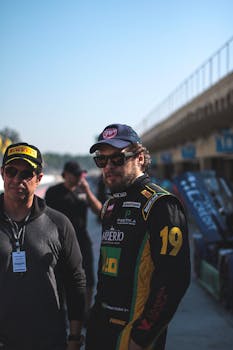 Two race car drivers in sunglasses and caps at a motorsport event, near racetrack.