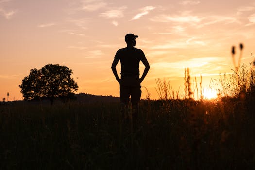 A man stands silhouetted against a vibrant sunset sky in a countryside meadow.