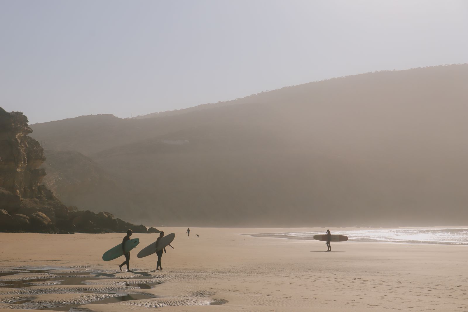 Surfers walking on a misty beach beneath cliffs on Portugal's surf coast