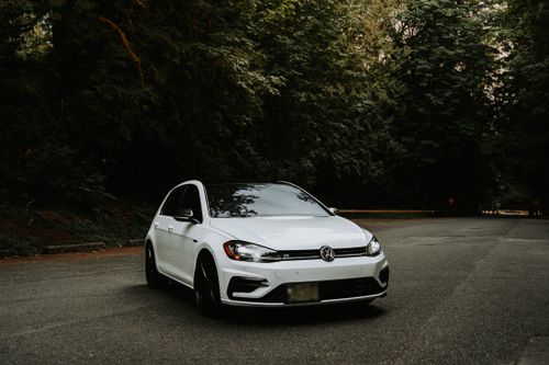 Free A white car with headlights on, parked on a forest road surrounded by lush green trees. Stock Photo