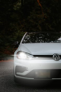 A close-up view of a white Volkswagen car with headlights on, set against a forest backdrop.