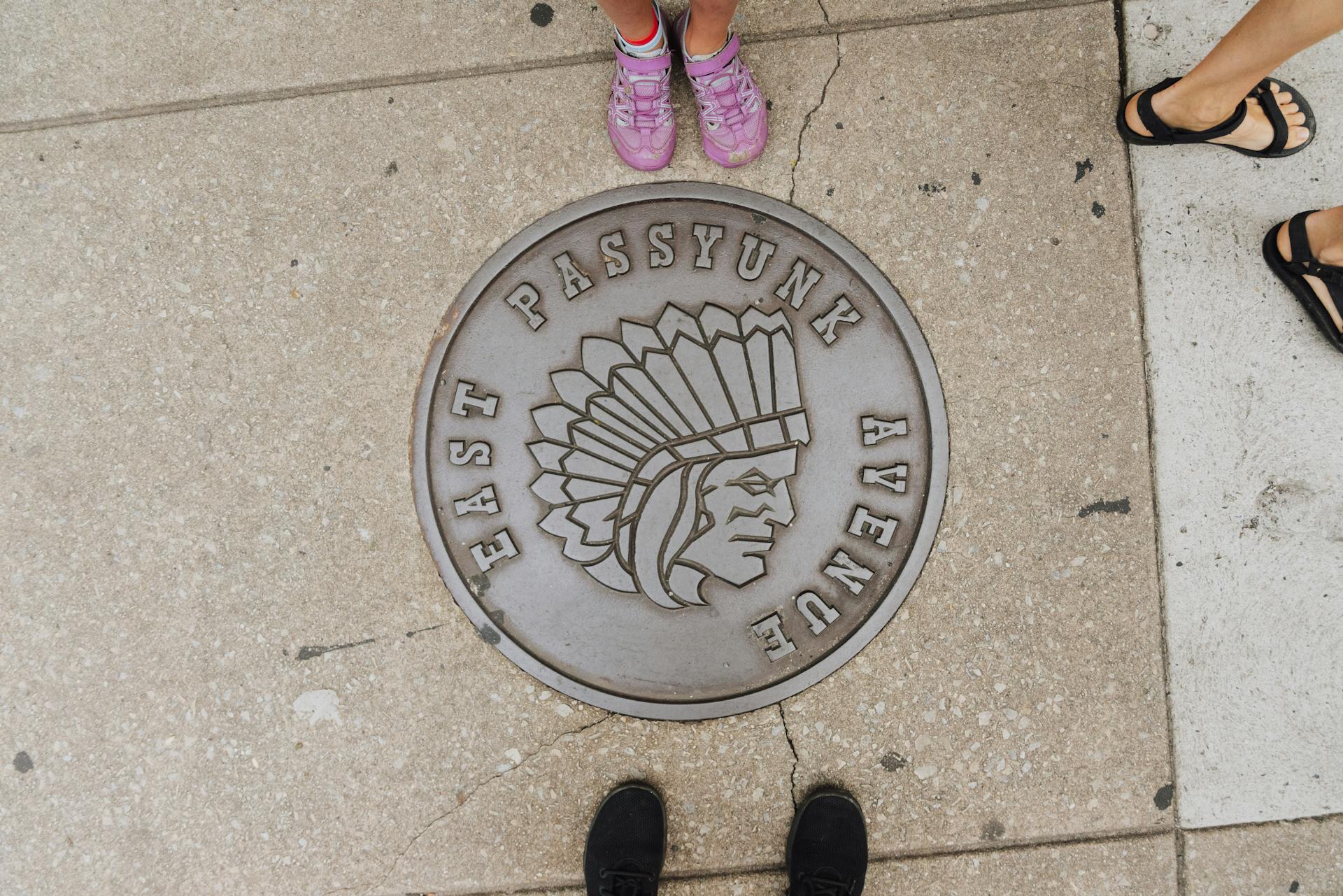 A metal manhole cover with a Native American design on East Passyunk Avenue sidewalk.