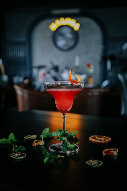 Free Vibrant cocktail with orange twist, garnished with mint, on a bar table with a blurred nightclub backdrop. Stock Photo