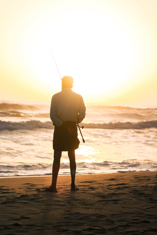 Back View of a Man Standing on a Beach at Sunset with a Fishing Rod 