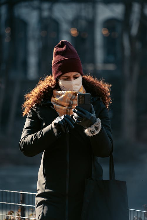 Woman in Coat with Face Mask Using Smartphone on Street