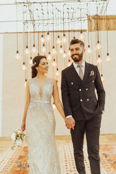 Smiling couple in elegant wedding attire holding hands under artistic lighting.