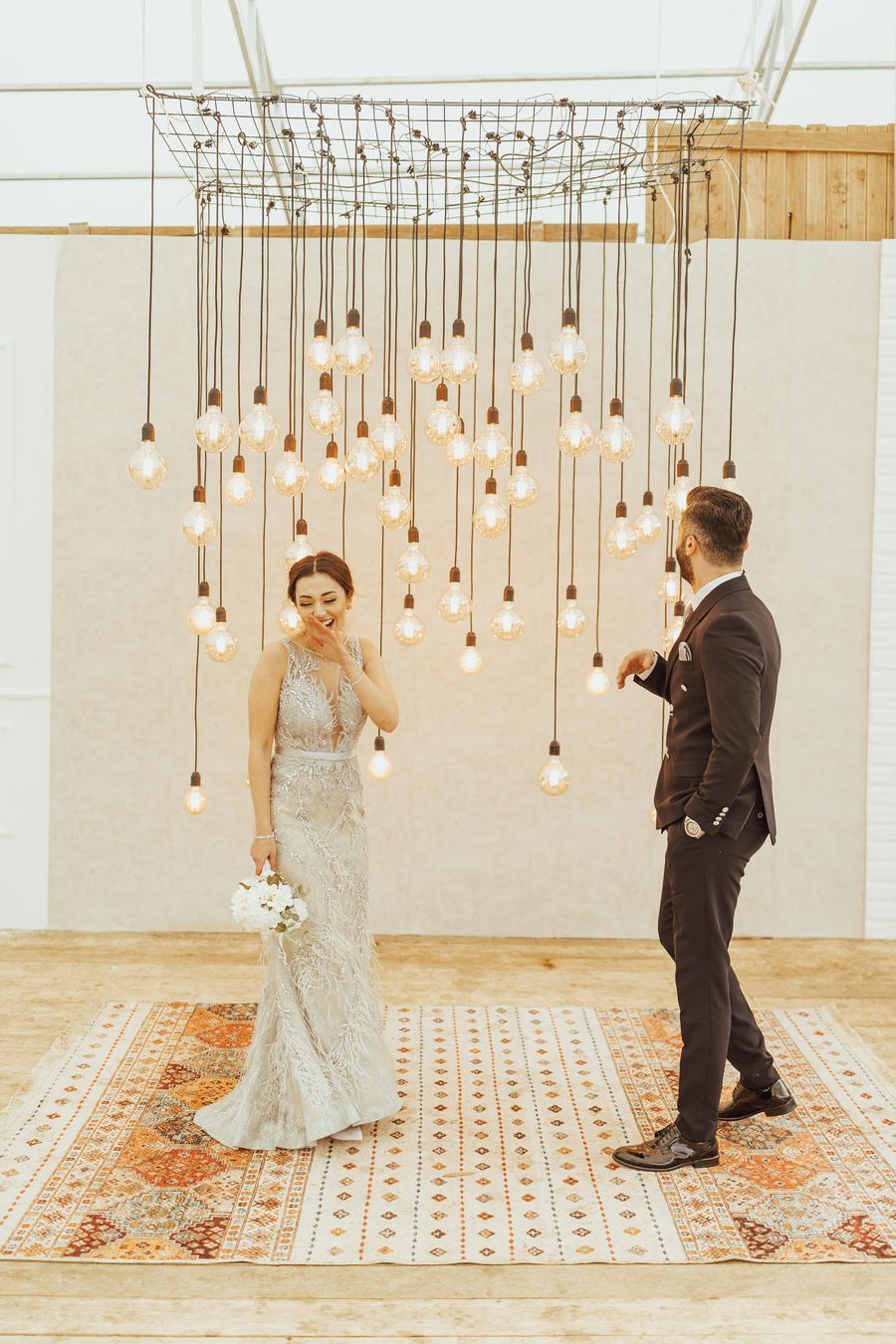 The couple standing beneath a cluster of hanging lamps on a patterned rug in a pale room