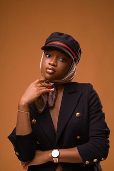 Stylish woman wearing a blazer, cap, and headscarf in a studio shot against a brown background.