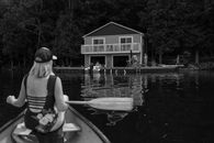 Woman paddling a canoe towards a lakeside house, capturing summer relaxation.