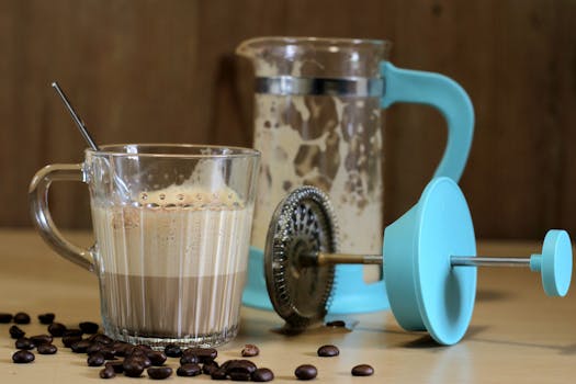 Glass coffee cup and blue French press with scattered beans on table.