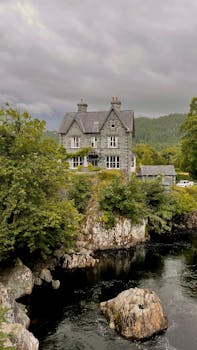 Elegant stone mansion amidst greenery by a serene river under overcast skies.