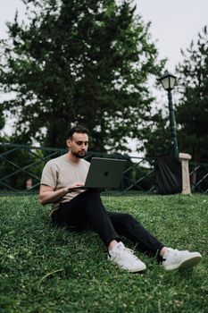 A young man sitting on grass using a laptop in a serene park setting, showcasing portable technology.