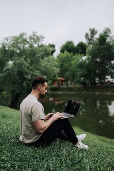 A man with short hair using a laptop while sitting on grass by a lake in a park.