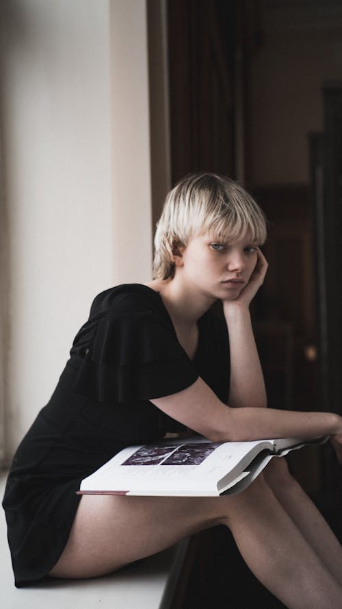 Free Young Blonde Woman Sitting on a Window Sill with an Art Book in her Lap Stock Photo