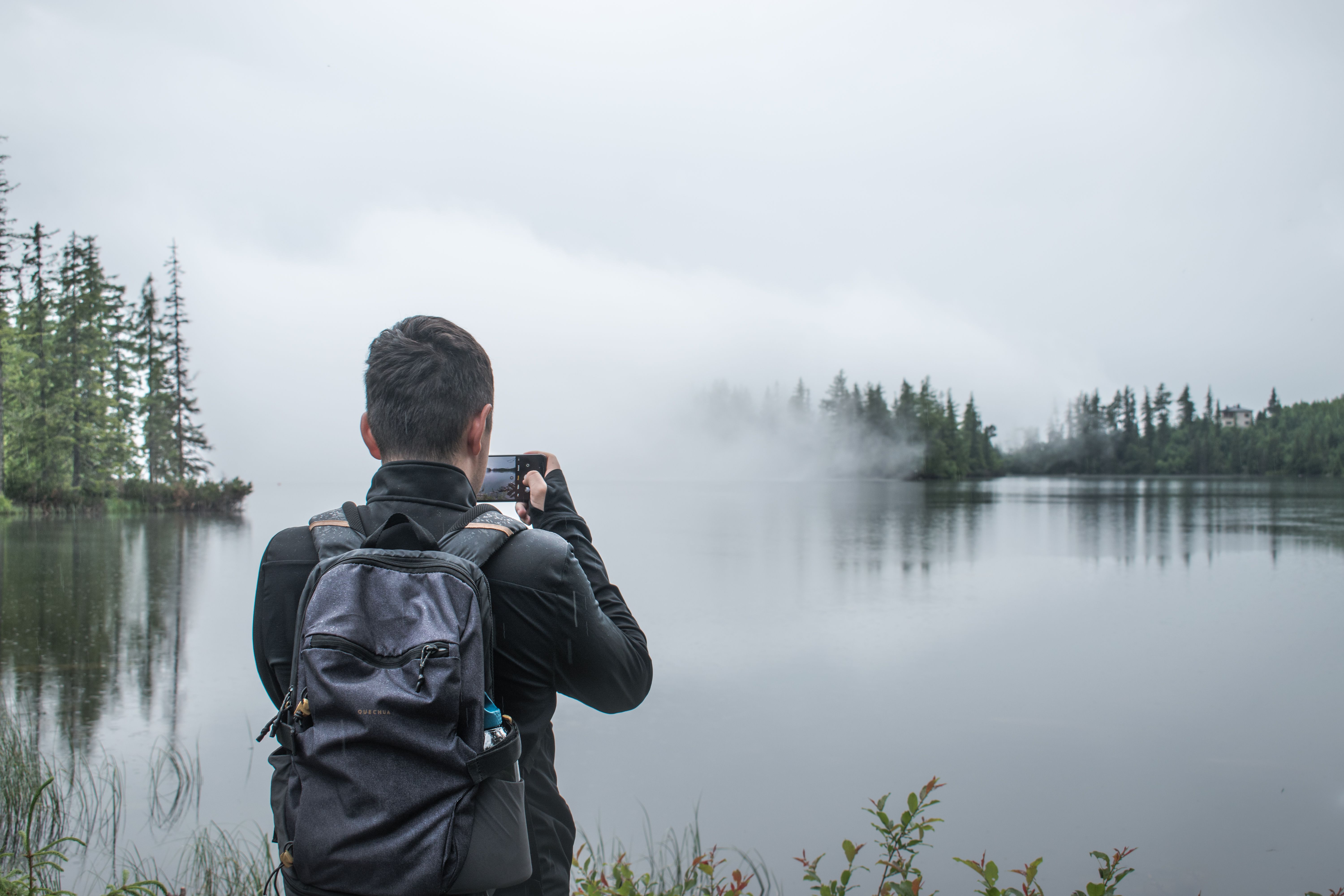 A man takes a photo of a misty lake scene with his smartphone, back to camera.
