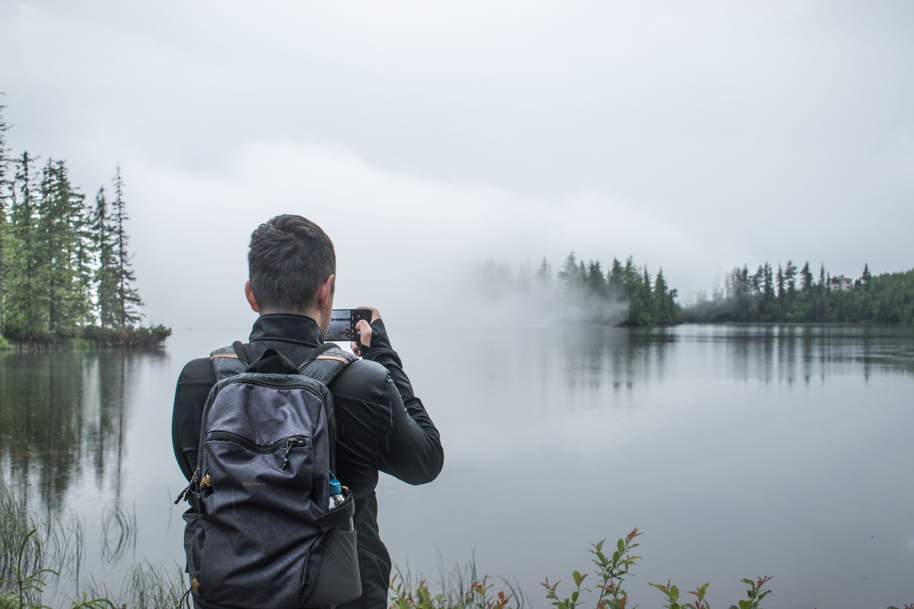 A man takes a photo of a misty lake scene with his smartphone, back to camera.