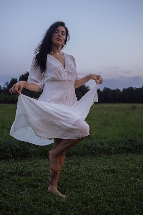 Young Woman in a White Dress Standing Barefoot on a Meadow 