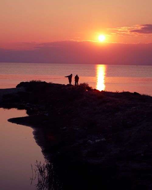Free Two silhouettes on a shoreline at sunset, reflecting serenity across the calm sea. Stock Photo