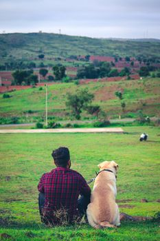 A man and his dog sit together overlooking a lush valley in Dharwad, India.