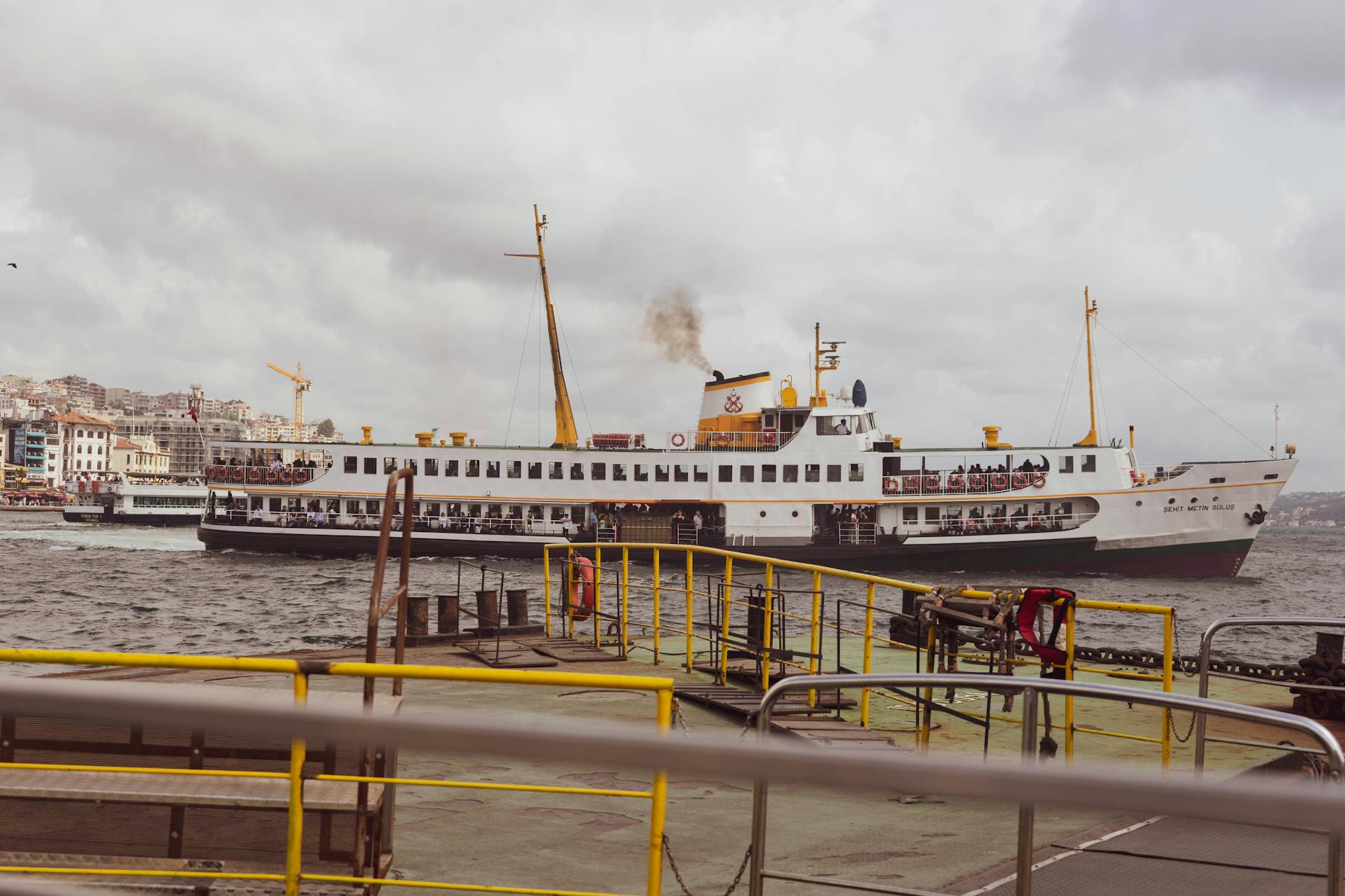 A ferry boat sails through a busy harbor, with an urban skyline in the background.