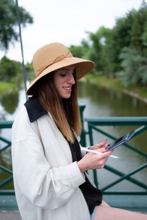 Free Smiling woman with long hair in a hat using a tablet outdoors. Stock Photo