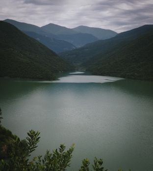 A tranquil mountain lake reflecting the surrounding lush green hills and cloudy sky.