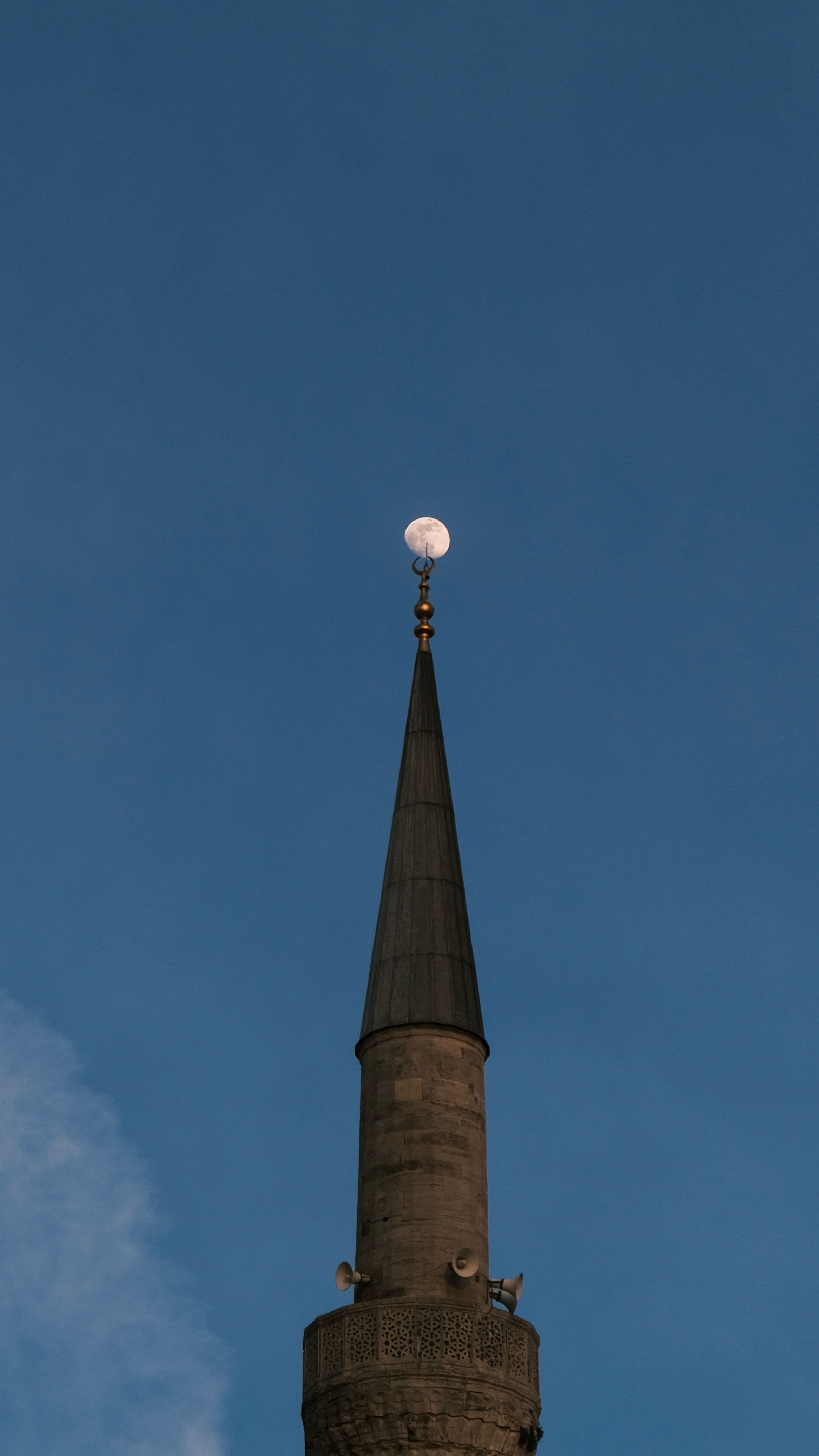 A striking view of a mosque minaret silhouetted against a clear evening sky with the moon overhead.