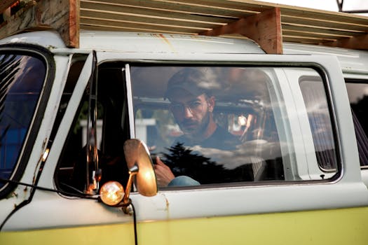 A man sits in an old van, seen through a window with reflections of trees and sky.