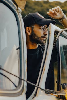A young man wearing glasses and a cap leans out of an old van window, gazing thoughtfully outdoors.